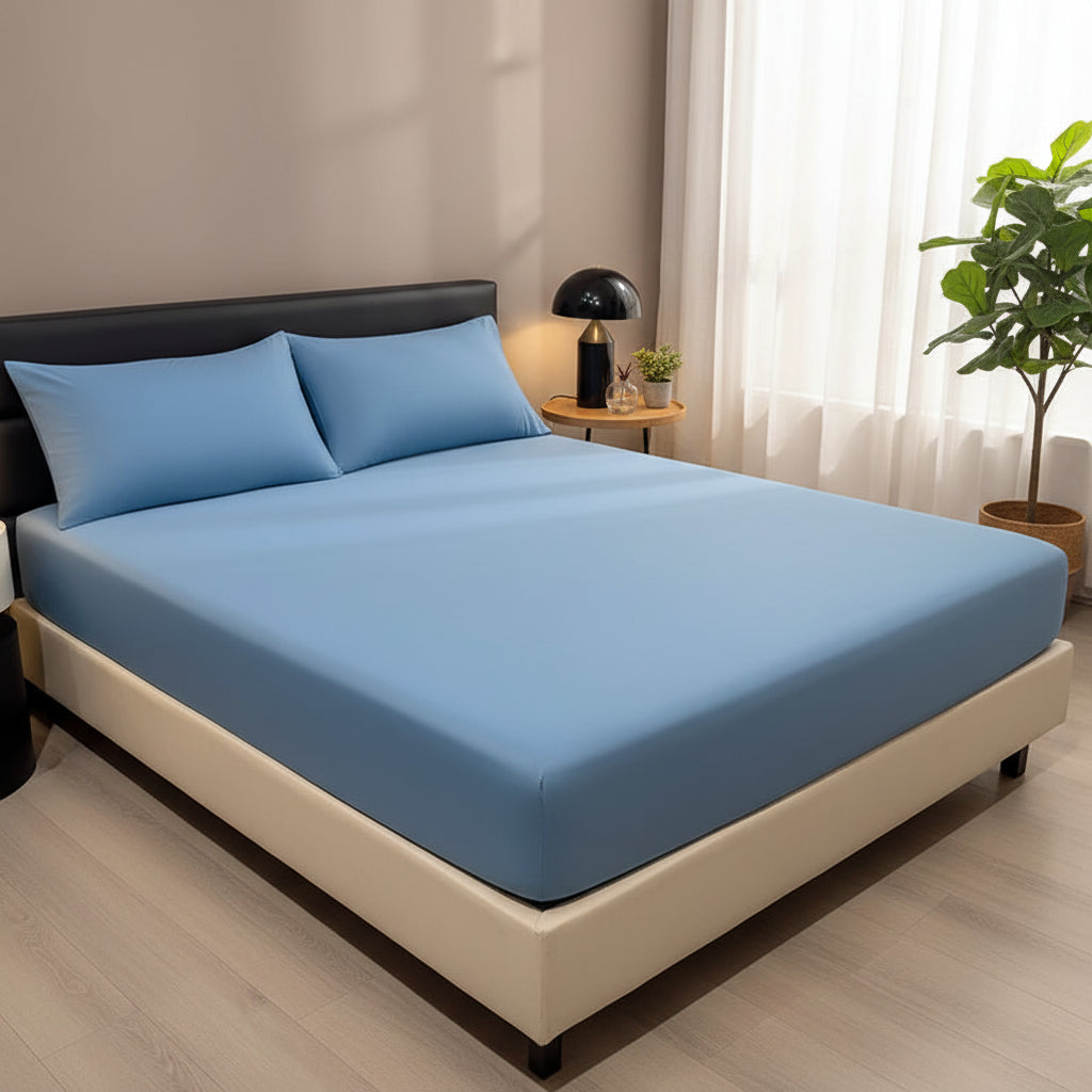 A light blue fitted bedsheet pulled tightly over a mattress corner by hands, shown on a bed in a bedroom setting. Two blue pillows and a decorative tray with a candle are also visible.